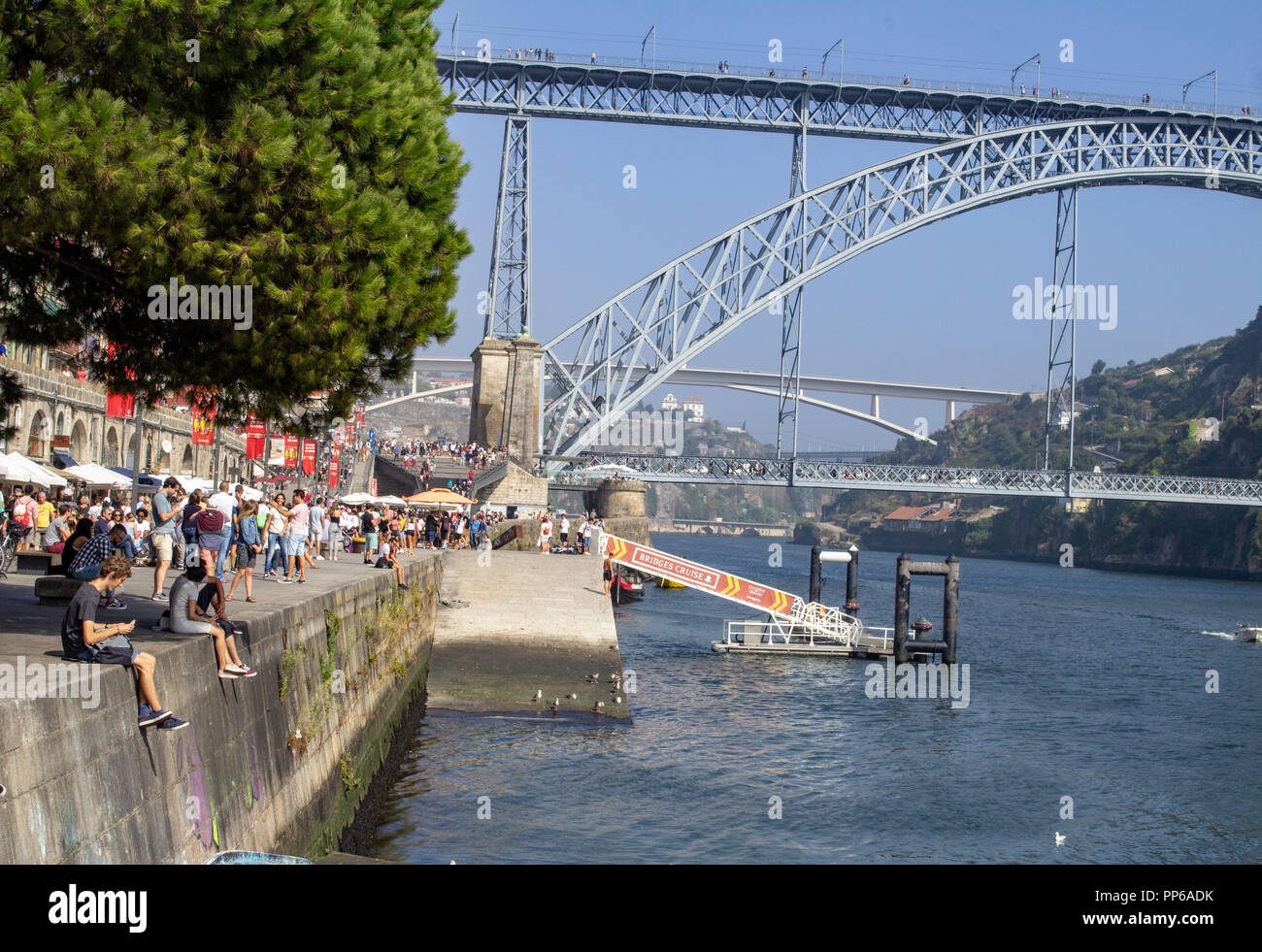 Porto, Portuga. Touristen drängen die Kais entlang des Flusses Duoro mit der Maria Pia Brücke im Hintergrund und einem kleinen Motorboot, das den Fluss segelt. Stockfoto