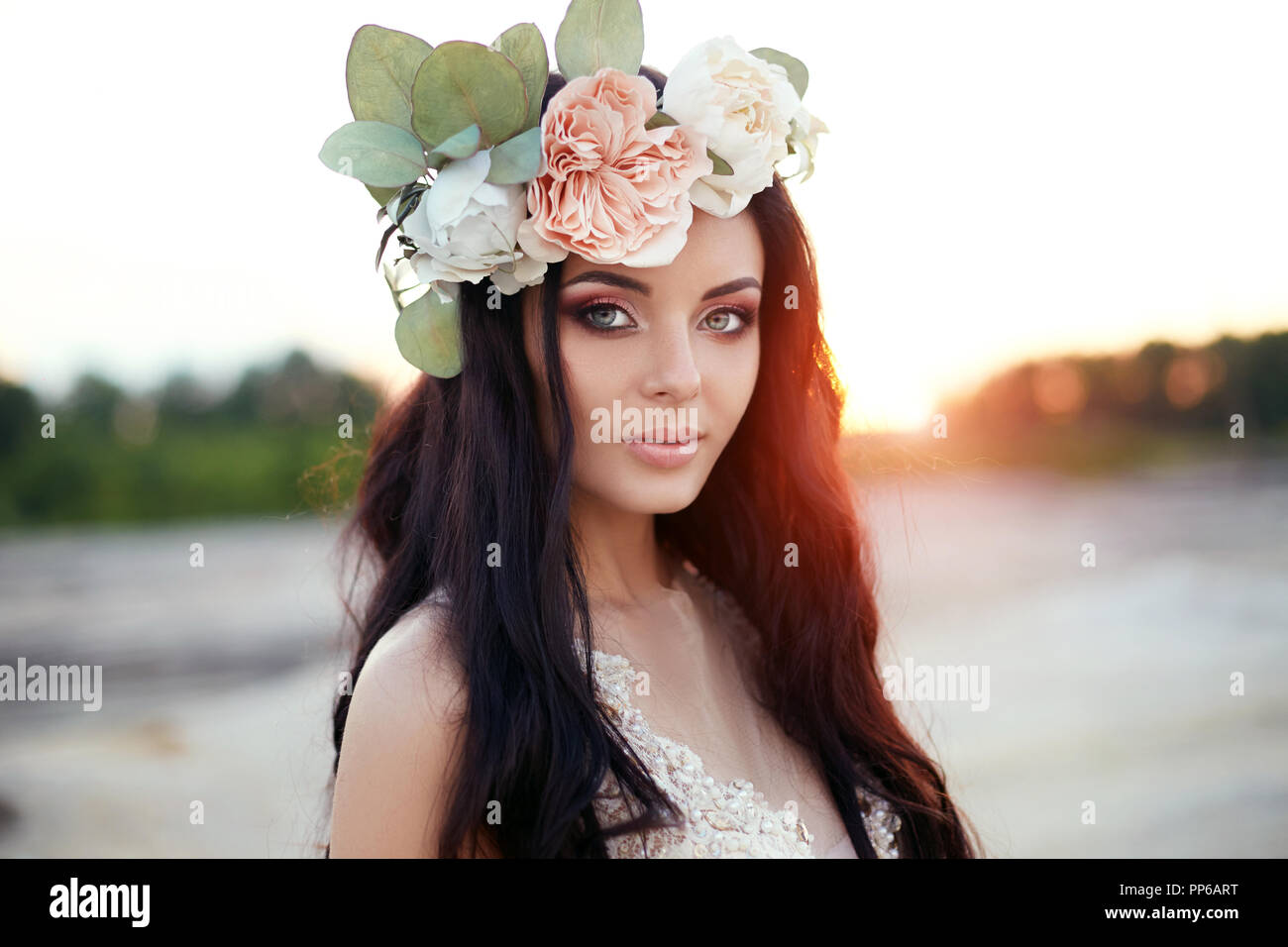 Frau mit einem Blumenkranz auf dem Kopf und einem Sommer Kleid Spaziergänge durch die Berge bei Sonnenuntergang. Porträt einer Brünette mit langen Haaren in den Strahlen der Sett Stockfotografie