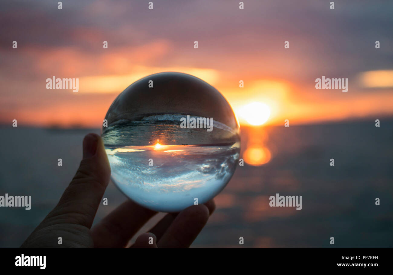 Helles Orange Marine Sonnenuntergang mit Welle und Sonne am Horizont gefangen in der Reflektion im Glas Kugel gehalten in den Fingerspitzen Stockfoto