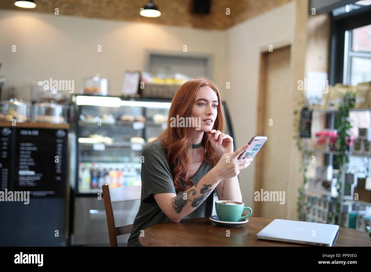 Red Head Mädchen mit Arm Tattoo, allein in einem Cafe an Ihr Mobiltelefon suchen sitzend, mit einem Laptop saß auf dem Holztisch in Liverpool, Großbritannien Stockfoto