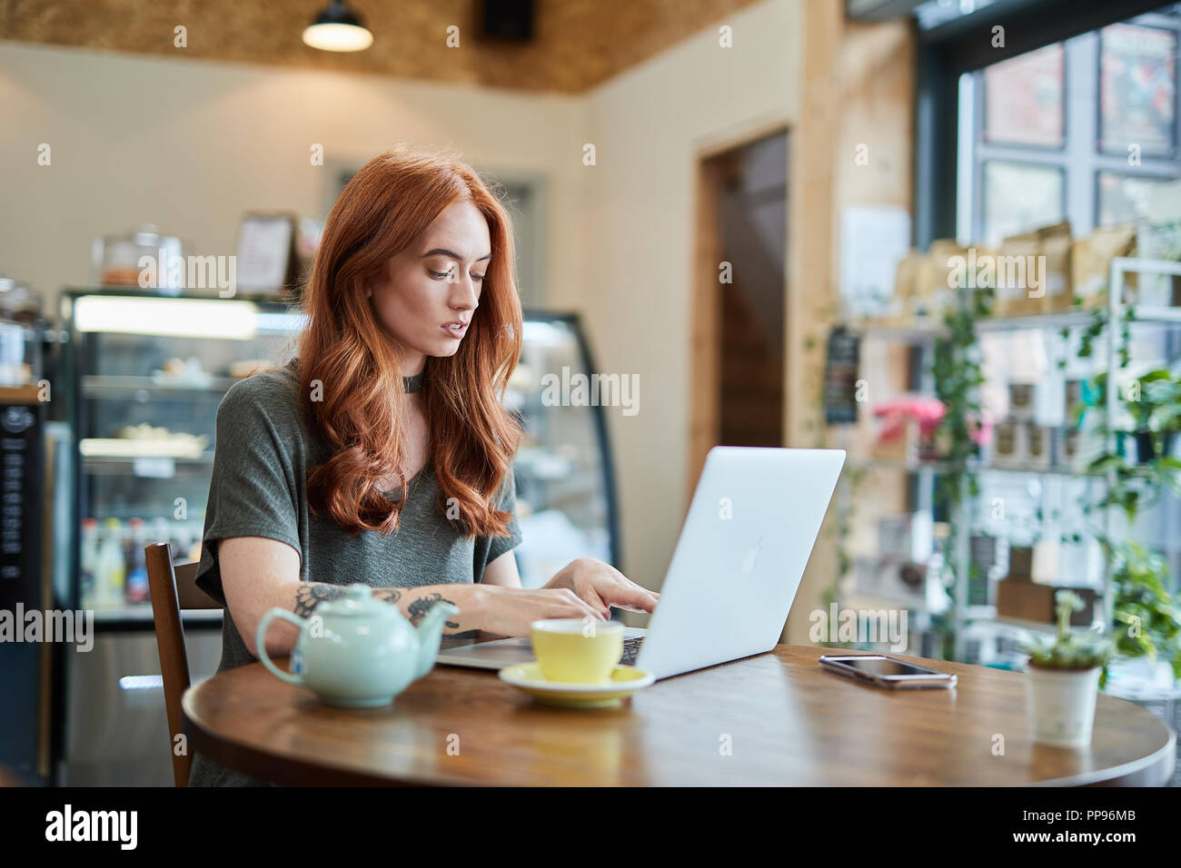 Red Head Mädchen mit Arm Tattoo, allein in einem Cafe, das Arbeiten mit einem Notebook, Handy und ein Laptop, Liverpool, Großbritannien Stockfoto