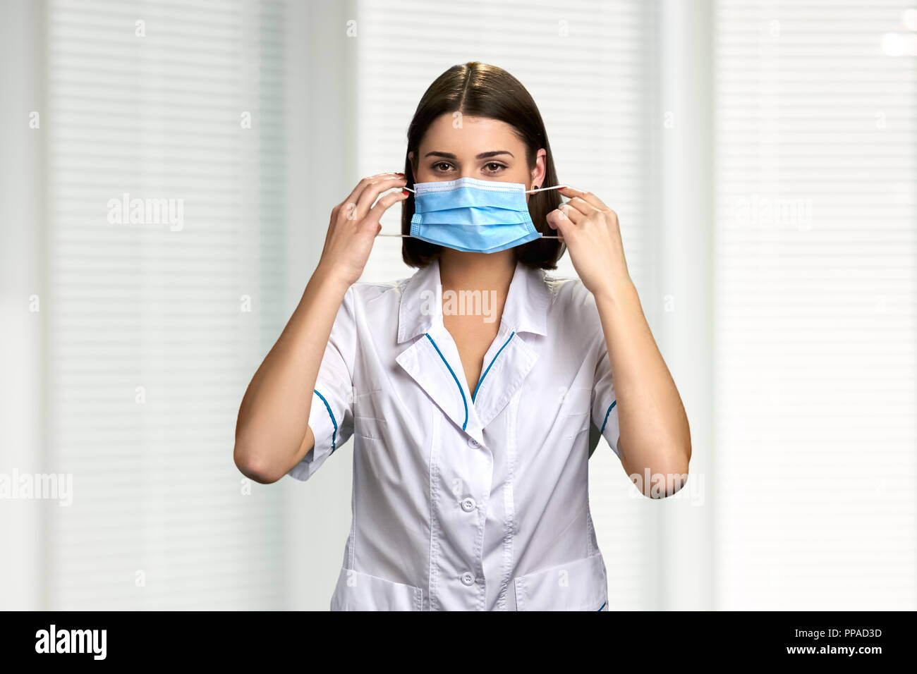 Ärztin auf die Gesichtsmaske. Junge brunette Arzt oder Krankenschwester im weißen Mantel setzt auf Gesichtsmaske im Krankenhaus Hintergrund. Stockfoto