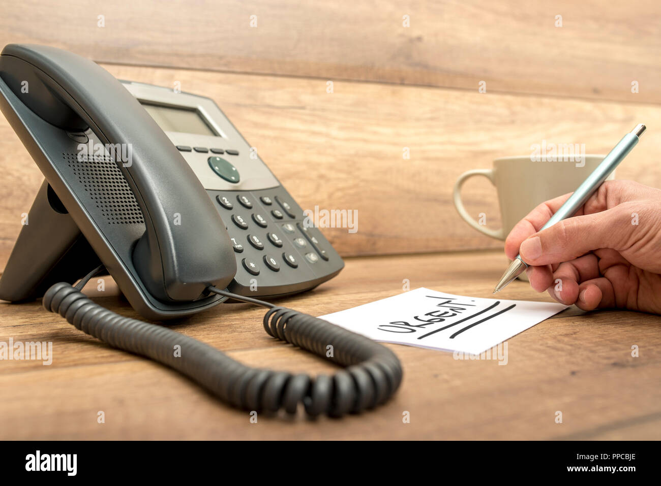 Nahaufnahme der männliche Mitarbeiter an der Rezeption schreiben das Wort dringend zweimal auf eine weiße Karte neben dem Festnetz Telefon unterstrichen. Stockfoto