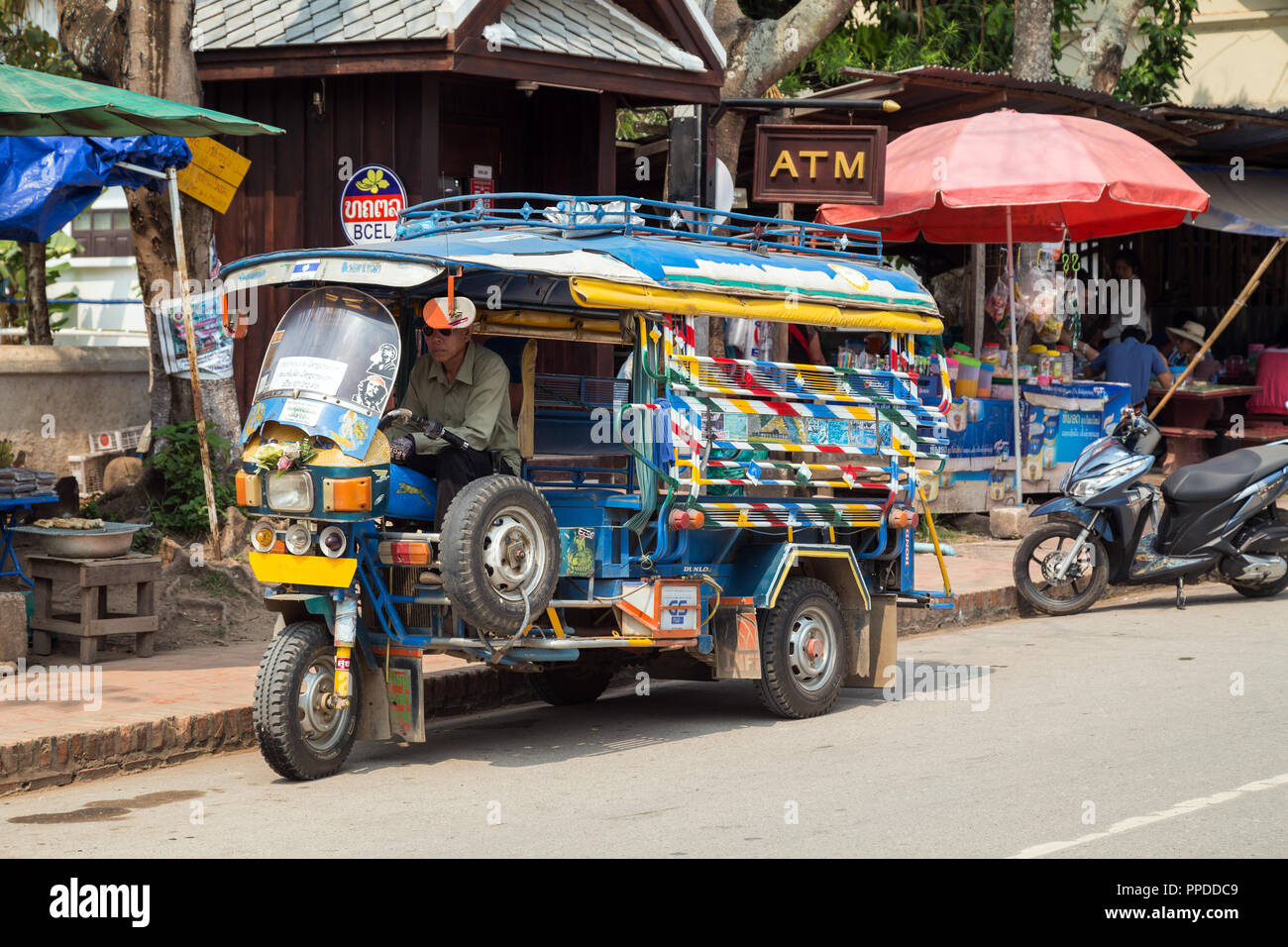 Treiber in einem geparkten bunten Dreirad Taxi genannt Jumbo (oder Tuk Tuk) auf dem Phetsarat Straße in Luang Prabang, Laos, an einem sonnigen Tag. Stockfoto