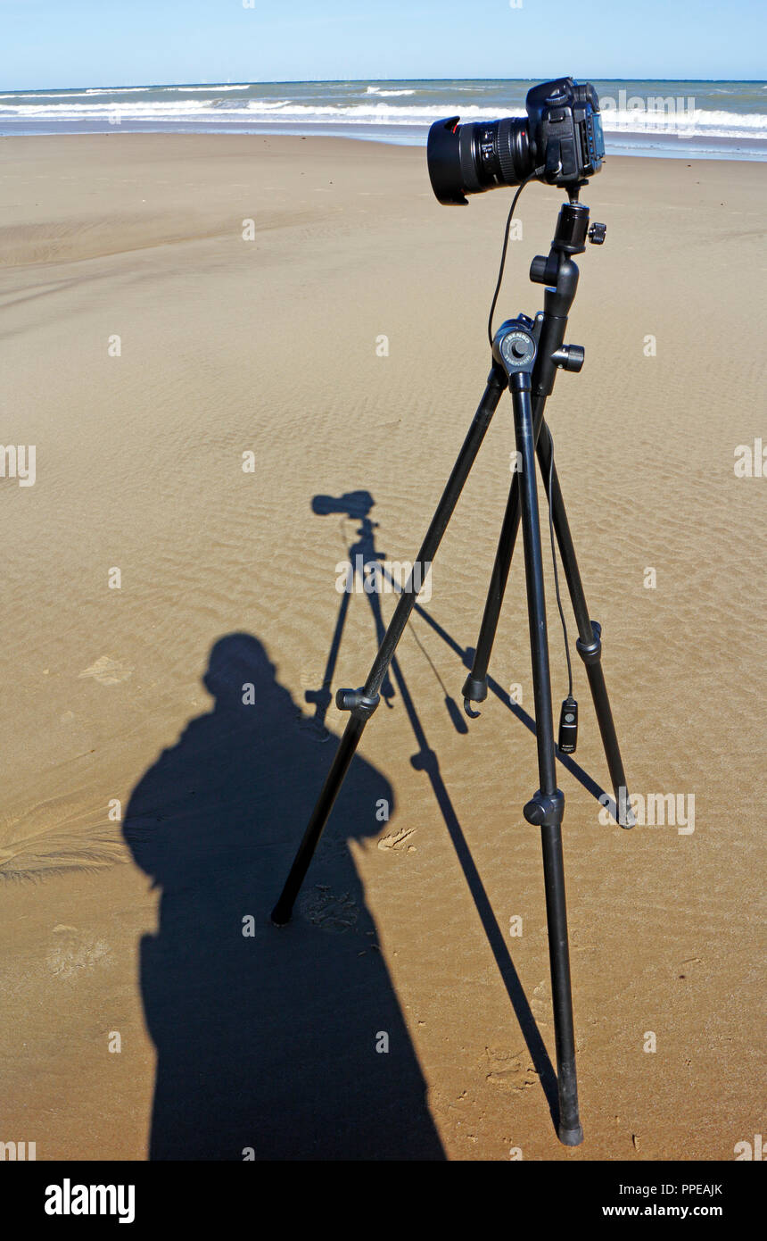 Eine Kamera auf Stativ mit Schatten einschließlich Fotograf an einem Sandstrand in North Norfolk, England, Vereinigtes Königreich, Europa. Stockfoto