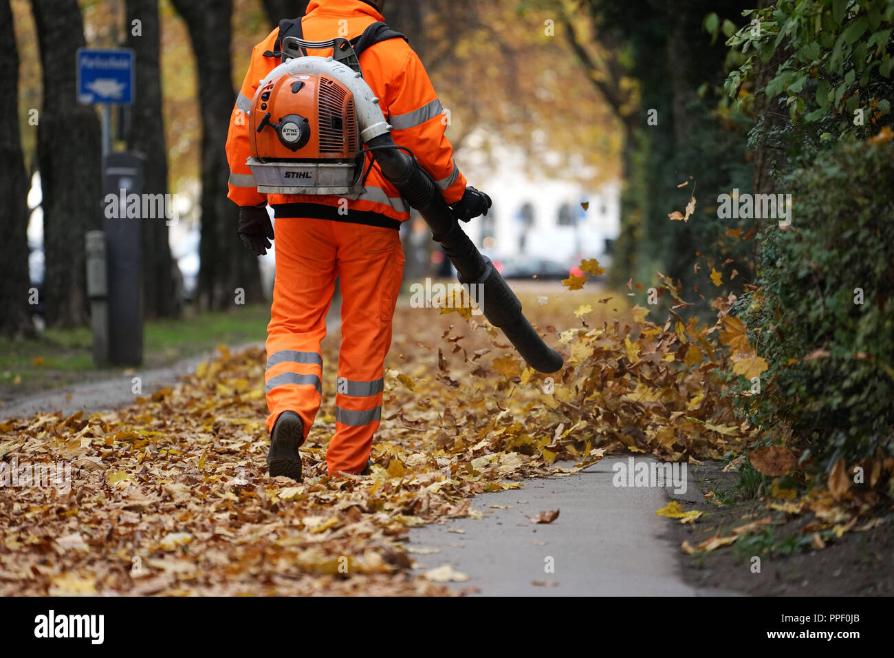 Ein Arbeiter in einem orange Overall bläst gefallen Blätter von einer öffentlichen Straße in München. Stockfoto