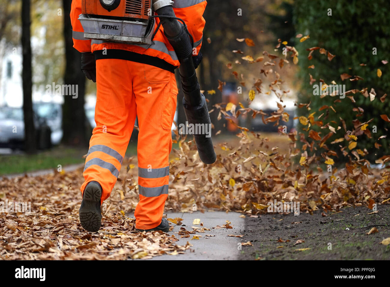 Ein Arbeiter in einem orange Overall bläst gefallen Blätter von einer öffentlichen Straße in München. Stockfoto