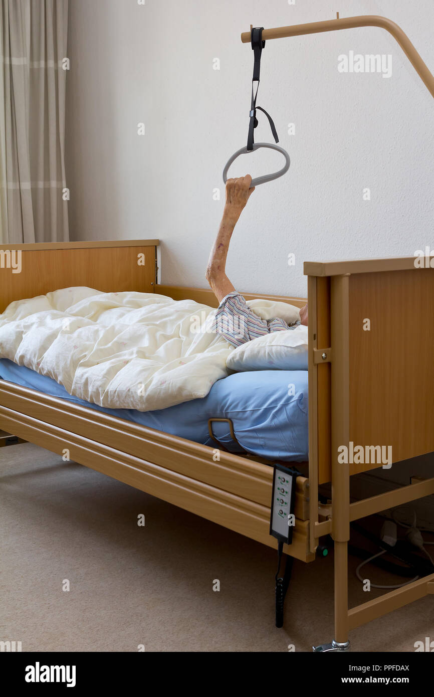 Alte Frau mit einer unheilbaren Krankheit, die versuchen, sich mit ihren extrem dünne und schwache Arme am Galgen der Krankenpflege Bett in ihrem Hospiz Zimmer zu ziehen Stockfoto