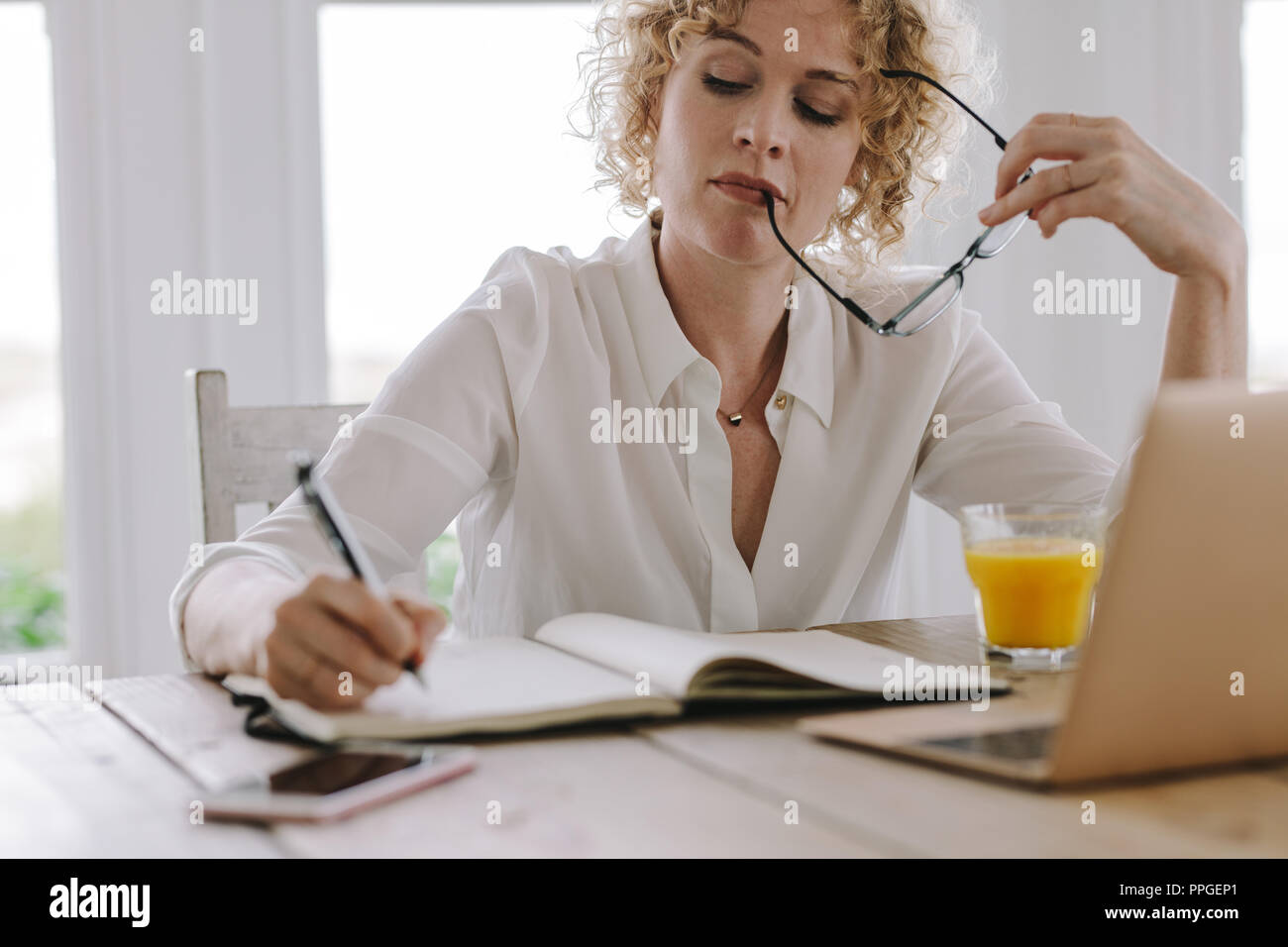 Frau von zu Hause aus arbeiten mit Laptop und Saft auf dem Tisch. Frau Verfassen von Notizen Tagebuch ihre Brille in der Hand halten. Stockfoto