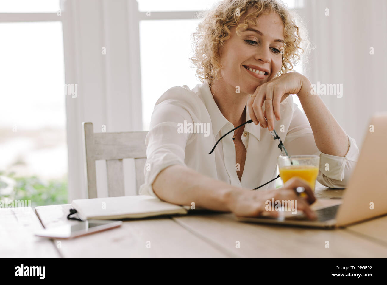 Lächelnde Frau von zu Hause aus arbeiten mit Laptop und Saft auf dem Tisch. Frau mit Laptop holding Brillen in der Hand. Stockfoto