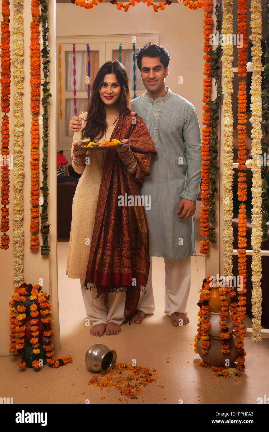 Glückliches junges Paar in traditioneller Kleidung steht in der Nähe der dekorierte Eingang ihres Hauses mit pooja Platte. Stockfoto