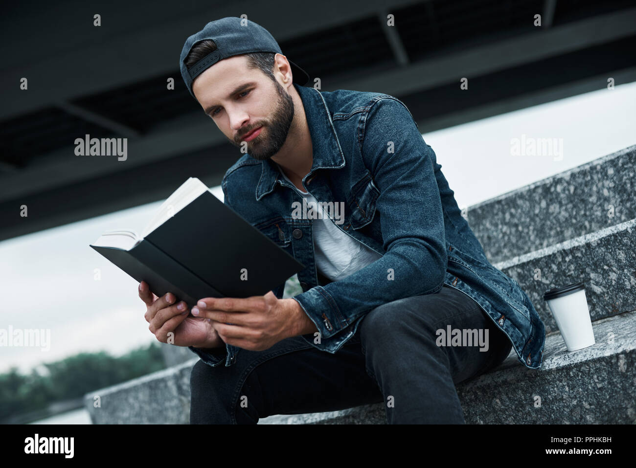 Freizeitaktivitäten im Freien. Junge stilvollen Mann sitzt auf der Treppe auf die Straße der Stadt bei einer Tasse heißen Kaffee beim Lesen Buch freudige close-up Stockfoto