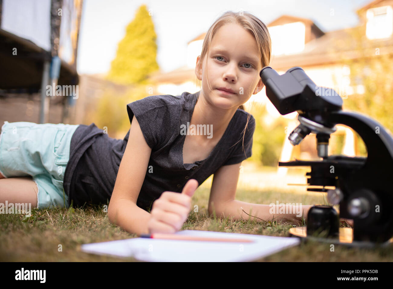 Ein 9 Jahre altes Mädchen ist, die auf dem Gras im Garten. Es hat ein Mikroskop und ein weißer Block vor. Das Mädchen sieht in die Kamera. Stockfoto