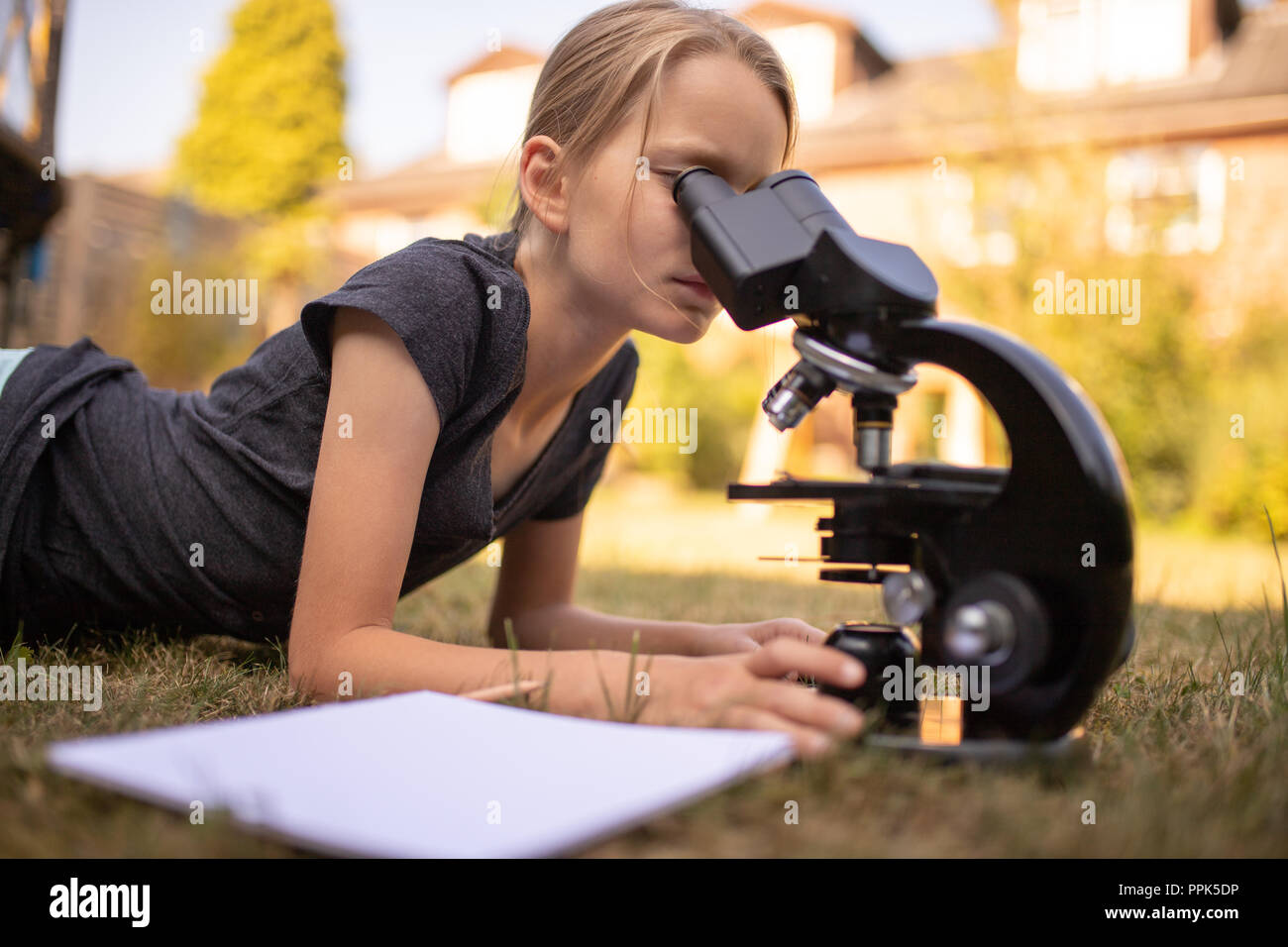 Ein 9 Jahre altes Mädchen liegt auf dem Rasen im Garten und schaut in das Okular eines Mikroskops. Vor Ihr ist ein Blatt Papier. Stockfoto