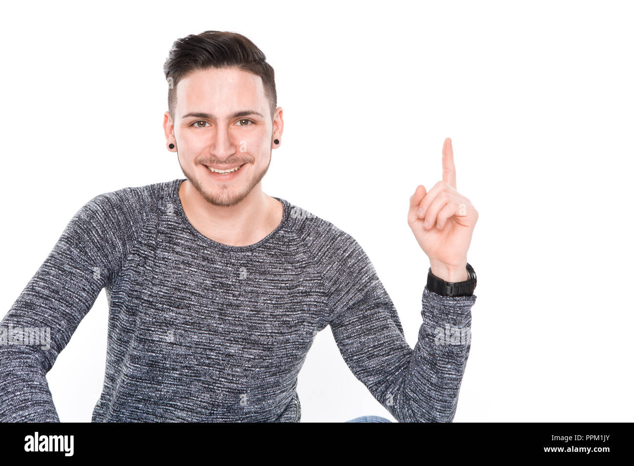 Junge brunette Mann mit Lächeln auf seinem Gesicht zeigen mit dem Finger auf etwas in high key Studio weißen Hintergrund zu Punkt Stockfoto