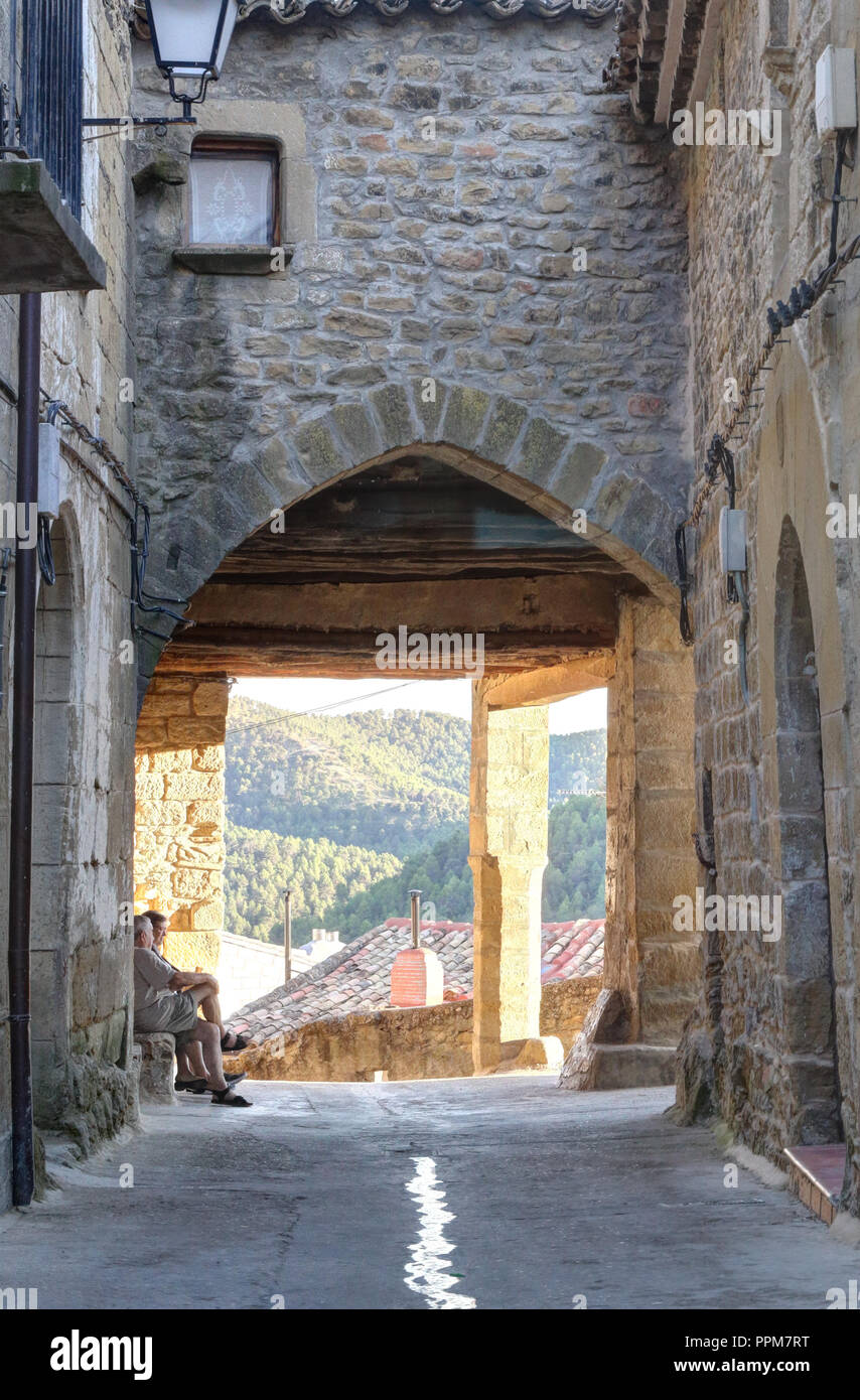 Zwei Männer bei Sonnenuntergang sitzen und plaudern zusammen unter einem Tudor Arch am Ende einer kleinen Stein Straße in der ländlichen Stadt Frago, Spanien. Stockfoto