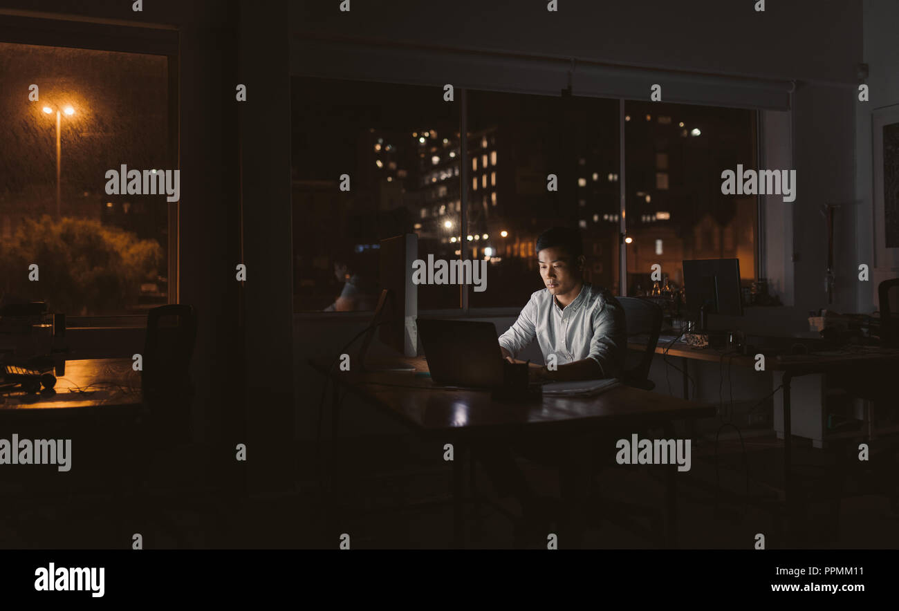 Junge asiatische Geschäftsmann an seinem Schreibtisch, die an einem Notebook arbeitet in einem dunklen Büro in der Nacht sitzen mit den Lichtern der Stadt im Hintergrund Stockfoto