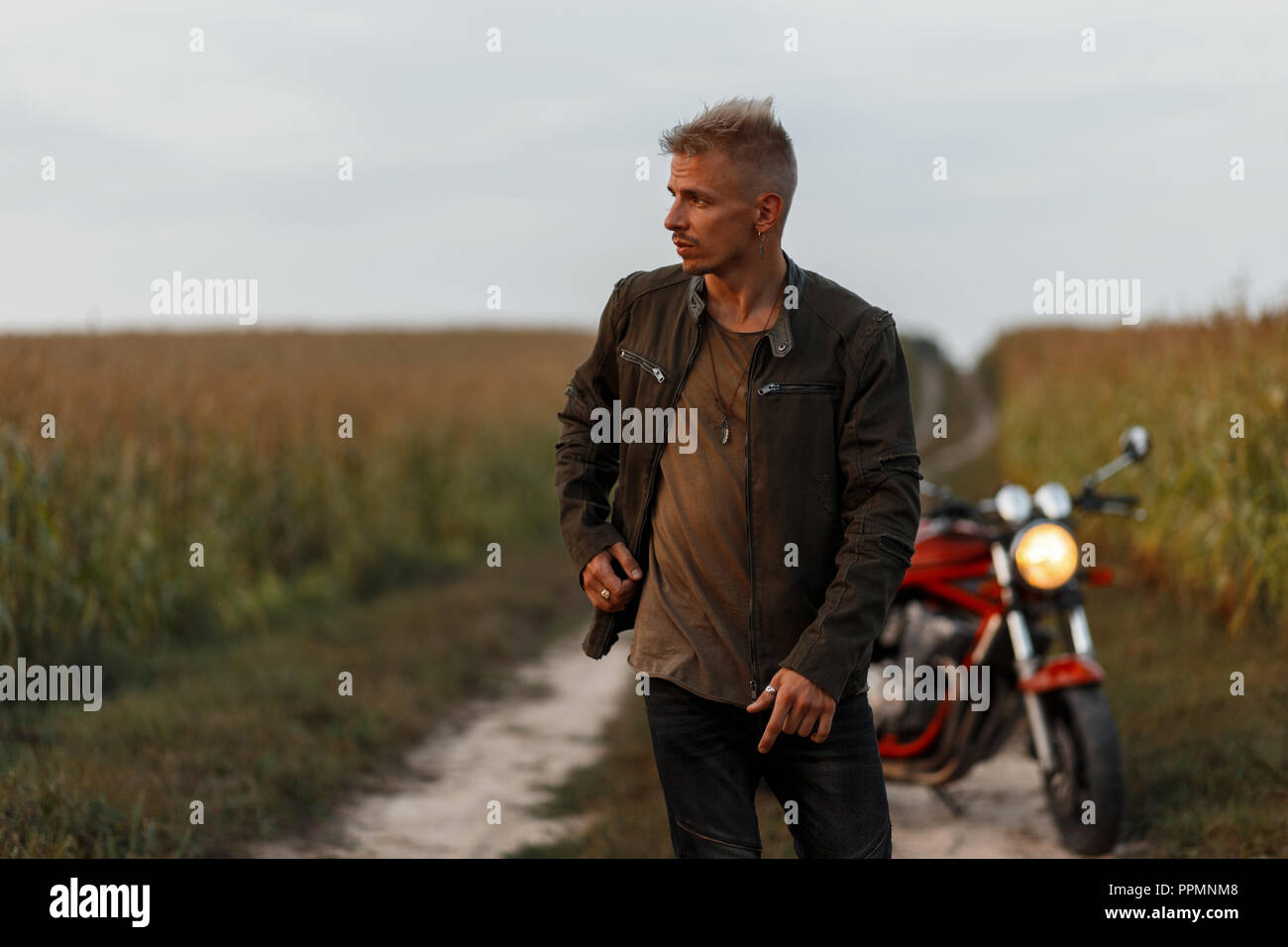 Stilvolle hübscher junger Modell Mann in einem khaki Jacke und T-Shirt mit einem Motorrad Reisen über die Natur in einem Maisfeld Stockfoto