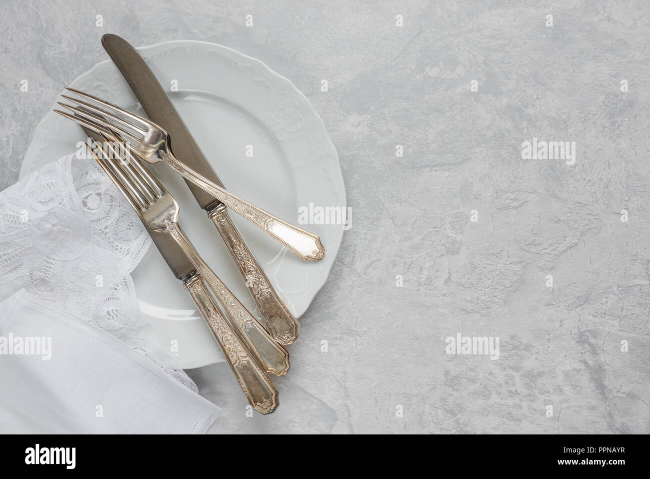 Silber Besteck auf einem Porzellanteller und weiß Serviette mit belgischen Spitze sind auf dem Hintergrund von grauem Beton Oberfläche, mit copy-Raum Stockfoto