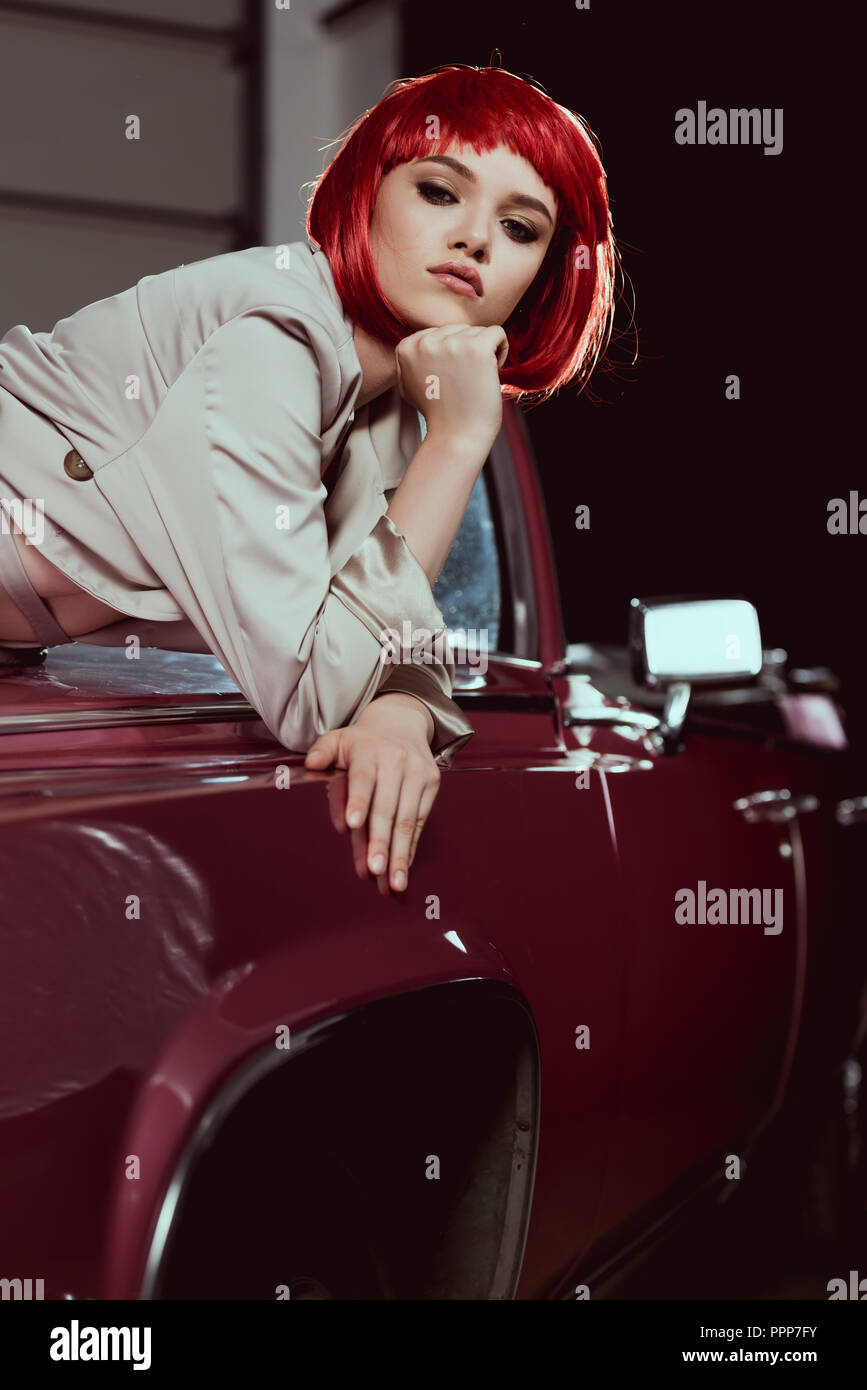 Attraktive junge Frau in Rot Perücke und Trenchcoat auf Kamera, während bei klassischen Auto gelehnt Stockfoto