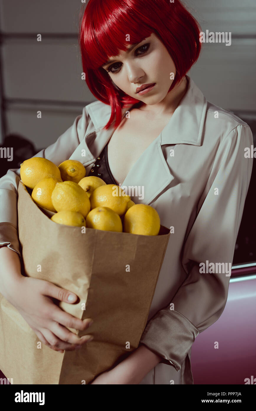 Nachdenkliches Mädchen in Rot Perücke und stilvolle Trenchcoat holding Paper Bag mit Zitronen Stockfoto