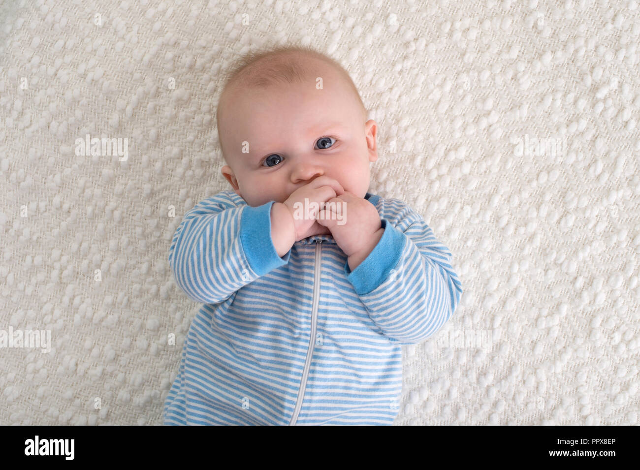 Ein 2 Monate altes Baby Junge lag auf dem Rücken auf einer weißen Decke. Er trägt eine blau-weiß gestreiften Schlafanzug und hat seine Hand in den Mund und Sucht Stockfoto