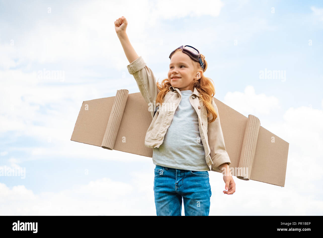 Porträt eines Kindes mit Paper Plane Flügel auf dem Rücken und schützende Brillen auf dem Kopf stehend mit ausgestrecktem Arm gegen den blauen bewölkten Himmel Stockfoto