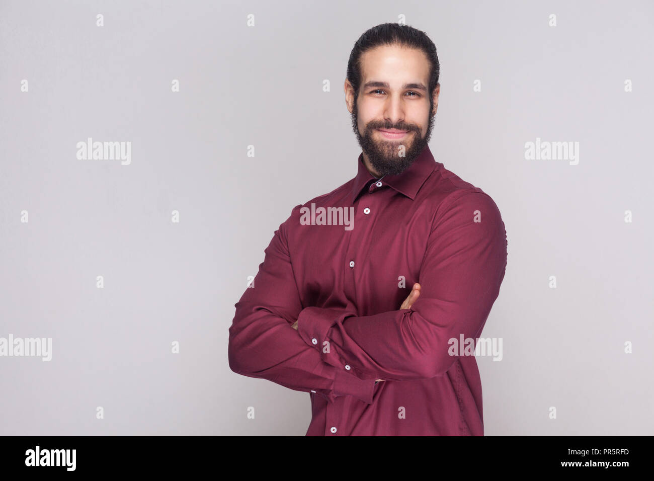 Portrait von stattlichen Geschäftsmann mit dunklem Haar und Bart im roten Hemd mit gekreuzten Händen, von der Kamera mit zufriedenen Gesicht und lächelt. Stockfoto