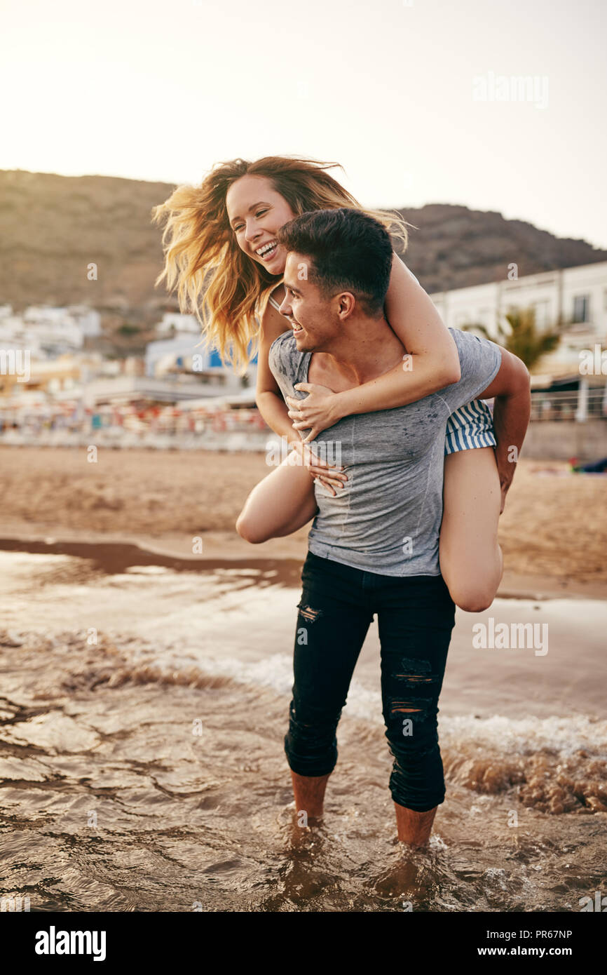 Lächelnden jungen Mann, der seine Freundin Lachen ein Huckepack in den Ozean und genießen Sie einen Tag am Strand Stockfoto