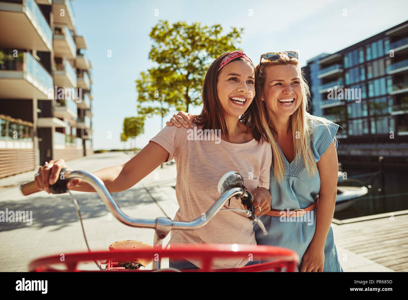 Zwei junge weibliche Freunde gehen Arm in Arm zusammen lachen mit dem Fahrrad durch die Stadt im Sommer Stockfoto