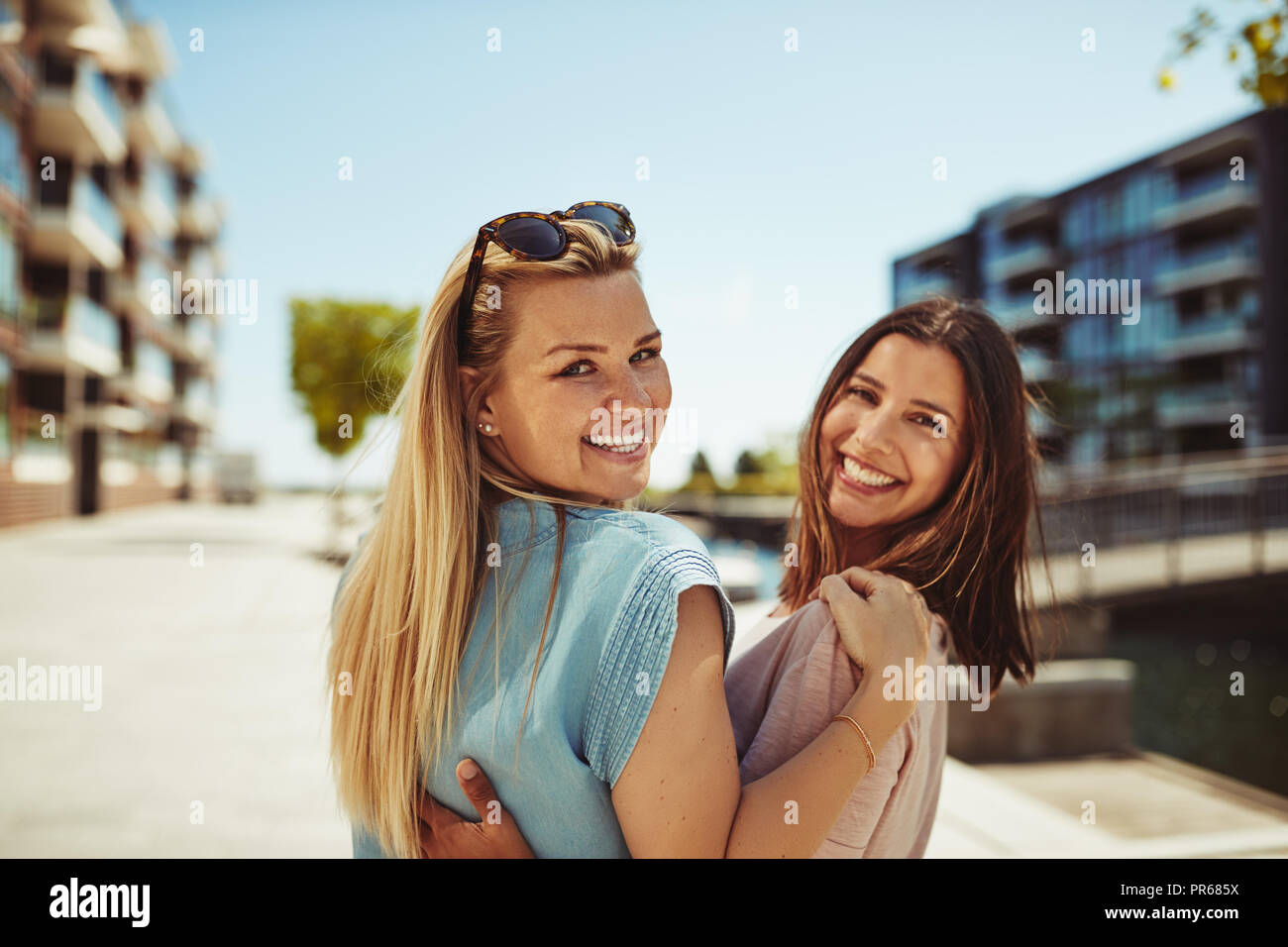 Zwei lachende Junge weibliche Freunde auf der Suche über die Schulter beim gehen Arm in Arm zusammen durch die Stadt im Sommer Stockfoto