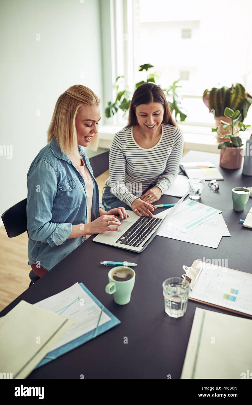 Zwei lächelnde junge Unternehmerinnen am Schreibtisch in einem Büro online arbeiten mit Laptop sitzen zusammen Stockfoto