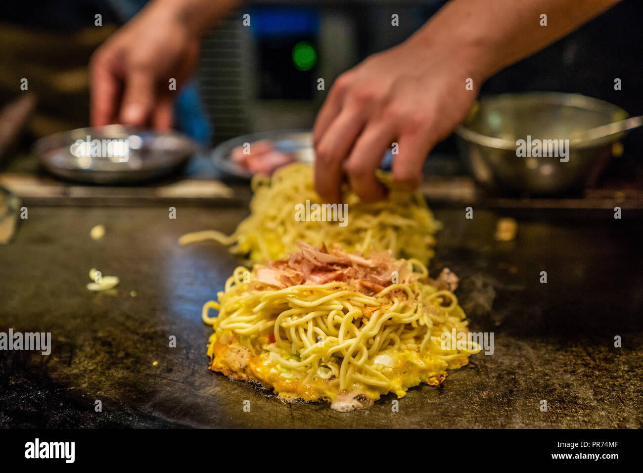 Chef kocht Okonomiyaki auf der Eisenplatte, er setzt die Nudeln in das Okonomiyaki, köstliche traditionelle Japanische Lebensmittel Stockfoto