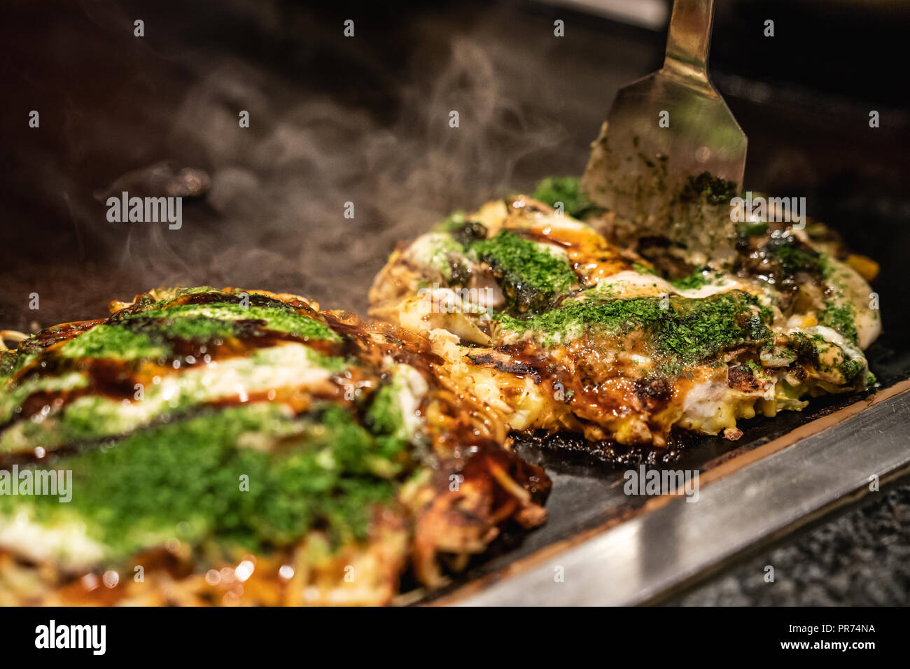 Koch mit Spachtel die getan okonomiyaki in der Hälfte auf der Eisenplatte zu schneiden, und bereit, die leckere traditionelle japanische Lebensmittel an den Kunden Stockfoto