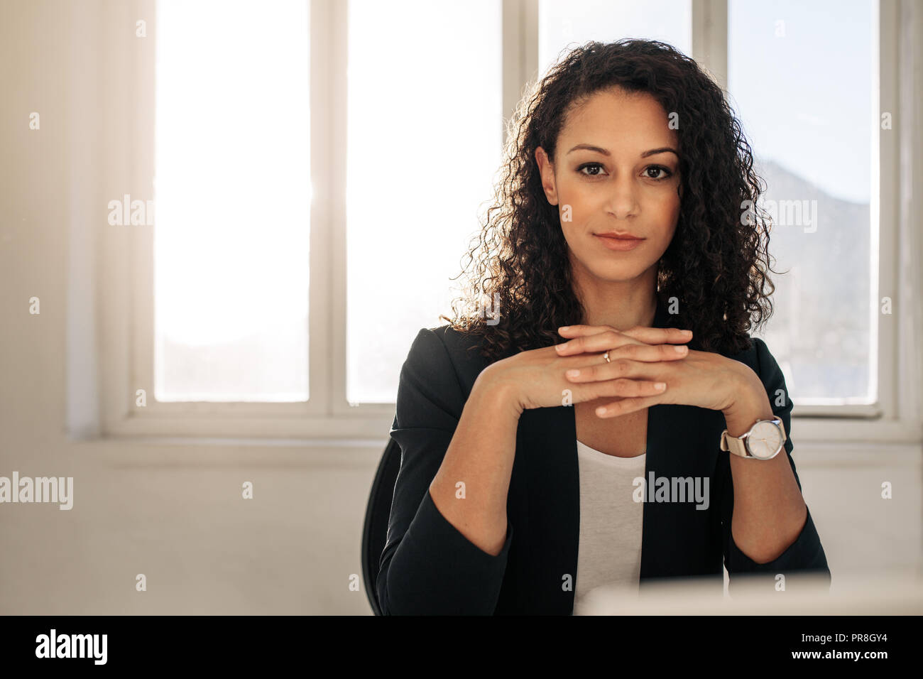 Geschäftsfrau, Sitzen an Ihrem Schreibtisch Verriegelung ihre Finger. Geschäftsfrau mit lockigem Haar in formelle Kleidung im Büro. Stockfoto