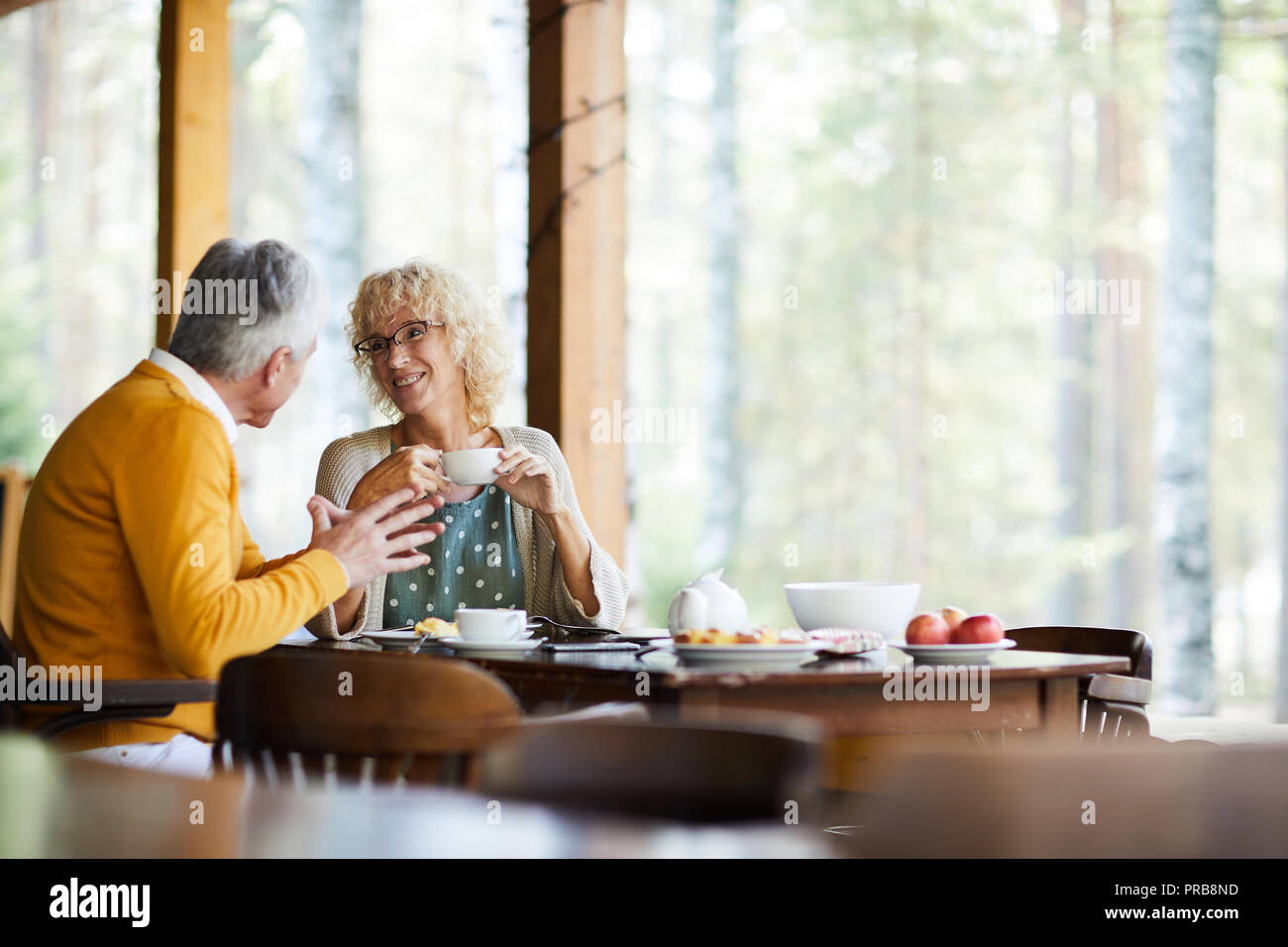Positive wunderschöne reife Paar in Freizeitkleidung sitzt am Tisch und trinken Kaffee im Veranda beim Chatten in ruhiger Ort Stockfoto