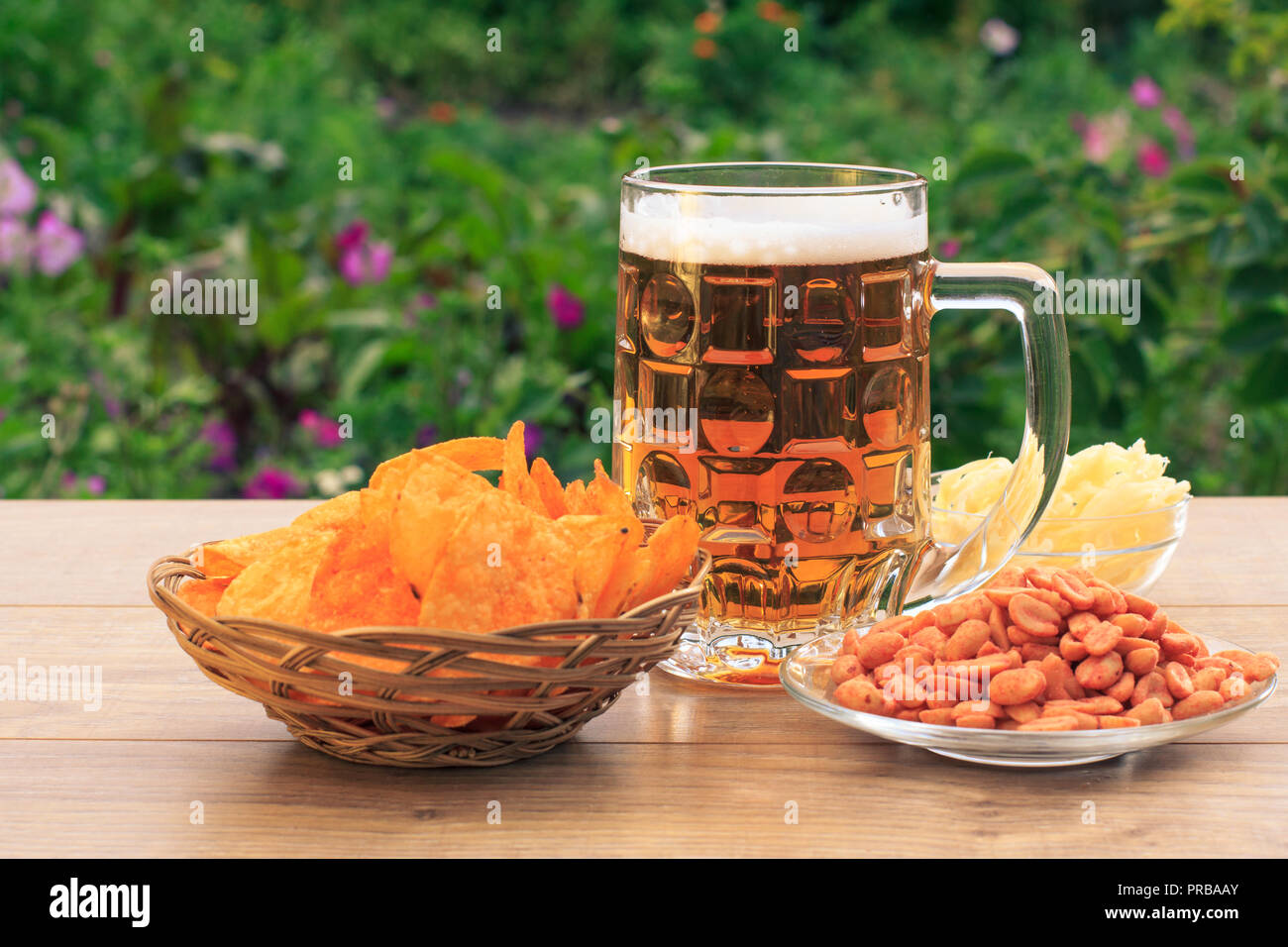 Glas Becher Bier auf Holztisch mit Kartoffelchips in Weidenkorb, Erdnüsse und getrockneten Tintenfisch in Schalen auf natürlichen, grünen Hintergrund Stockfoto