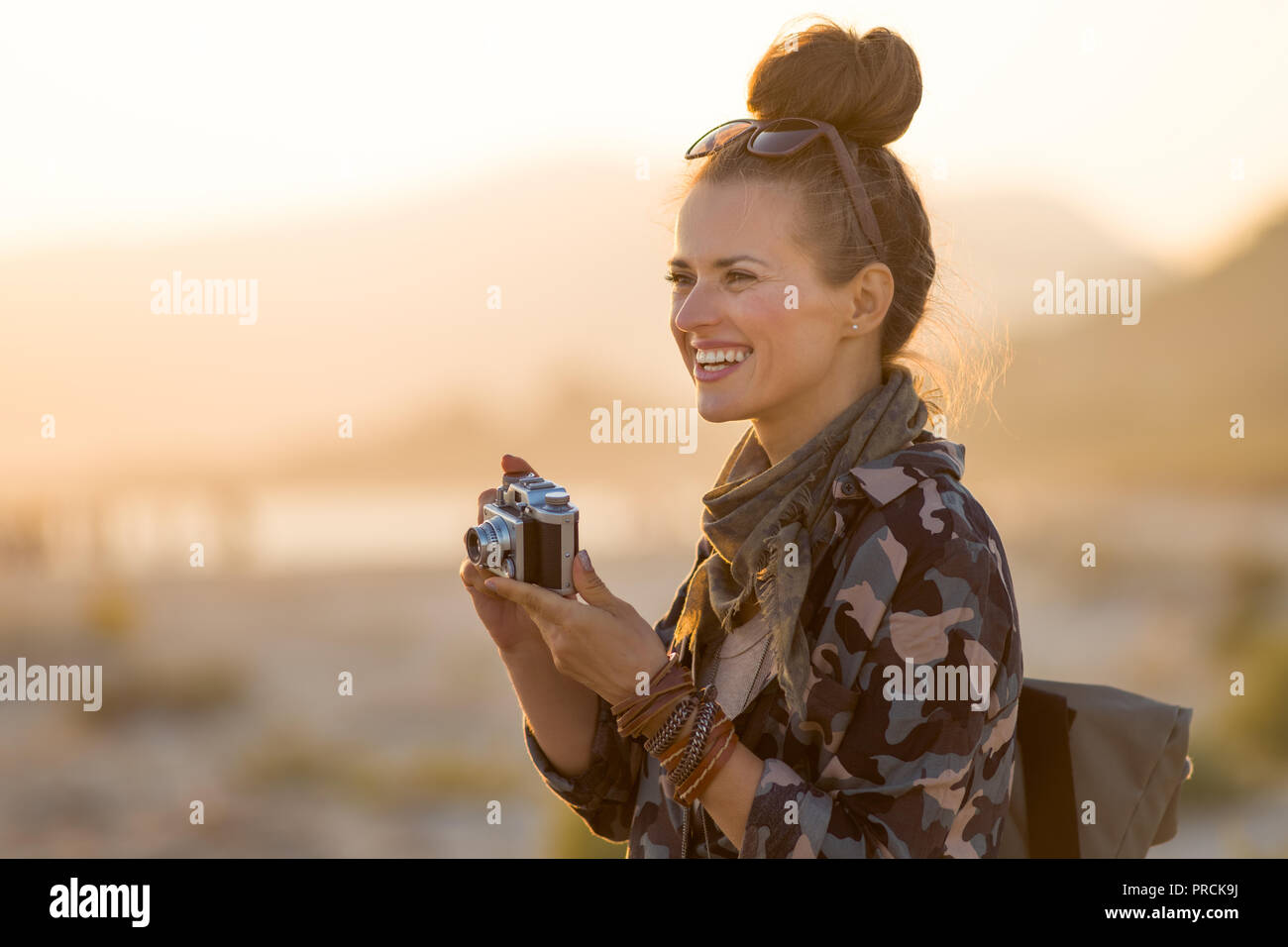 Glückliche junge Frau in Wanderkleidung mit retro Foto Kamera gegen Berg und Meer Landschaft bei Sonnenuntergang Stockfoto