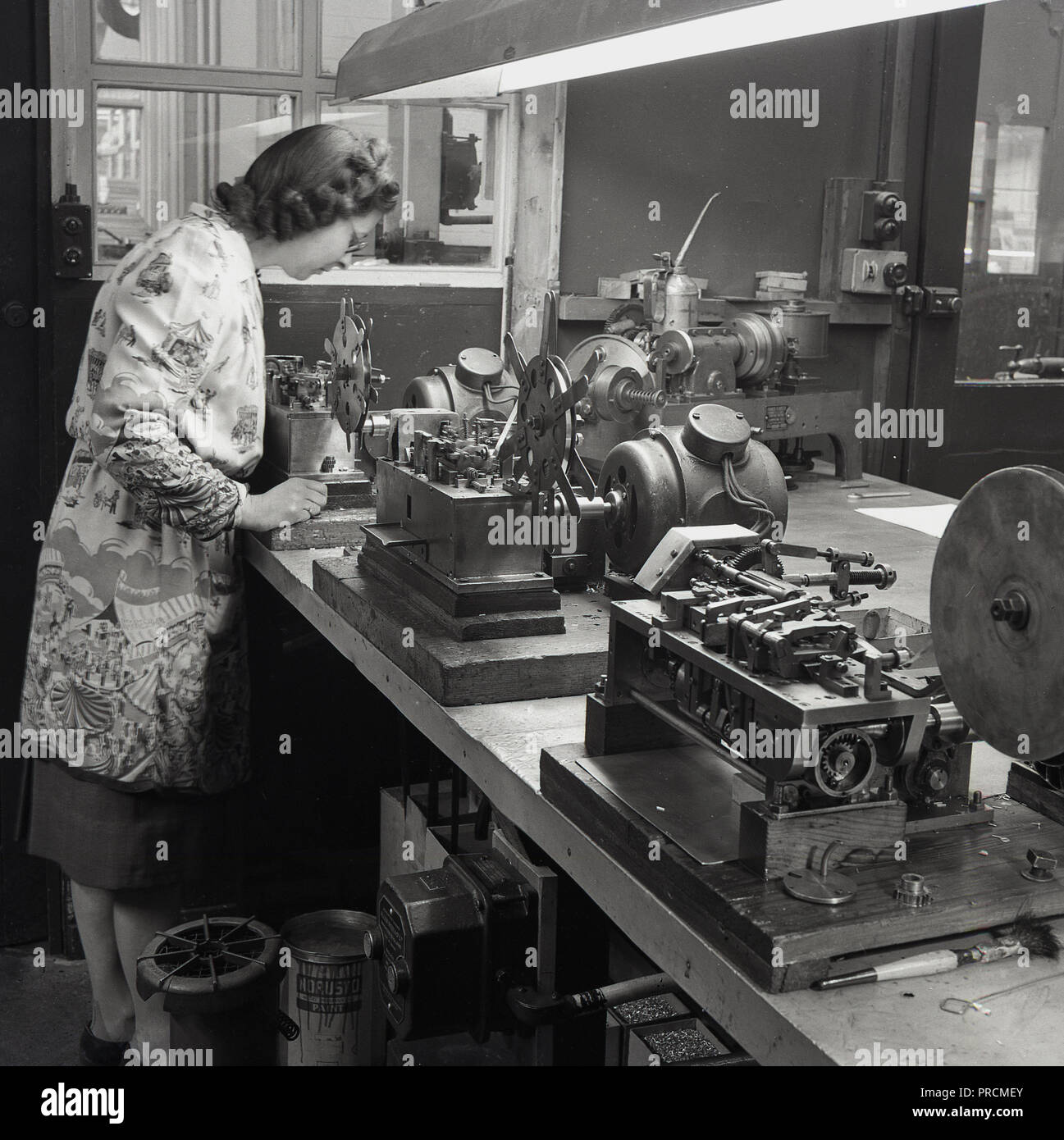 1950, historische, eine Dame Arbeiter in einer langen Ärmeln Schürze Schürze in einer industriellen Workshop des EFR, stehend auf einer Werkbank erfolgen, die die Kontrolle über die einzelnen Precision Engineering Werkzeugmaschinen auf die Bank über Holz- Grundlagen fest. Stockfoto