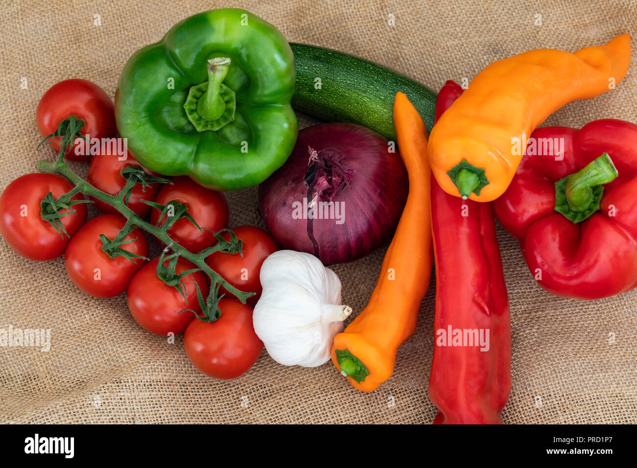 Nahaufnahme von roten, grünen und orangen Paprika und Paprika. Weintomaten, Knoblauch, eine rote Zwiebel und eine Zucchini. Gemüse auf einem hessischen Tuch. Stockfoto