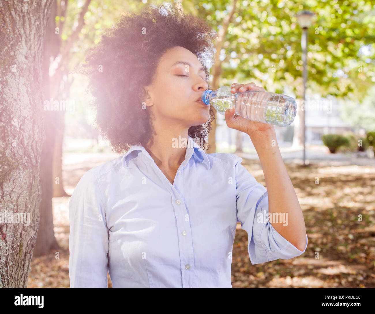 Schöne junge afrikanische Frau trinkt Wasser aus der Flasche und Entspannung in der Natur, Verschleiß legere Kleidung, Sonnenlicht flare im Hintergrund Stockfoto