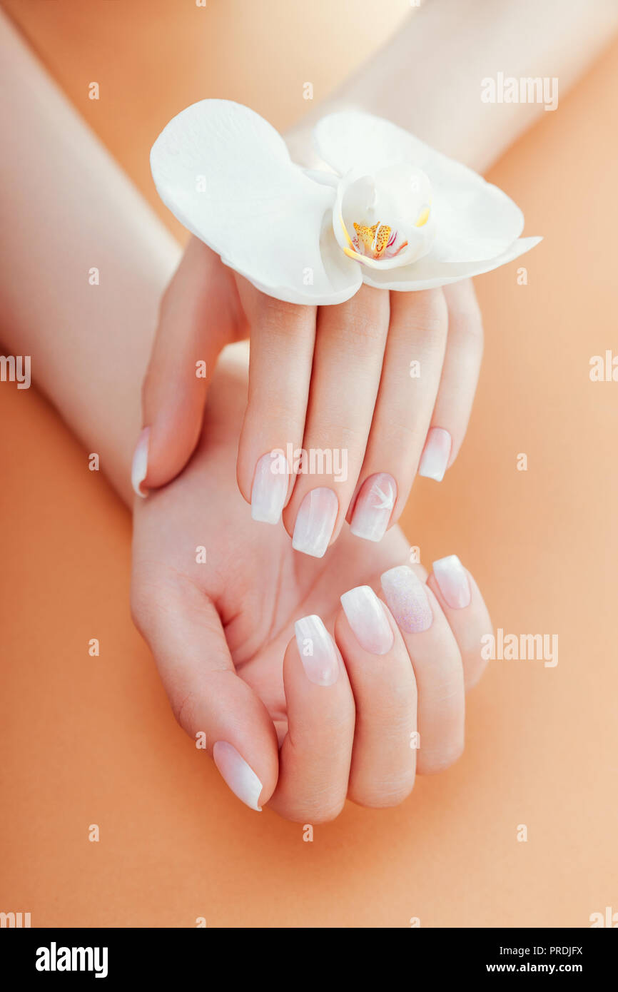 Ombre French Maniküre mit funkelt und Orchidee auf orangem Hintergrund. Frau mit weißen ombre French Maniküre hält Orchideenblüte. Körperpflege Stockfoto