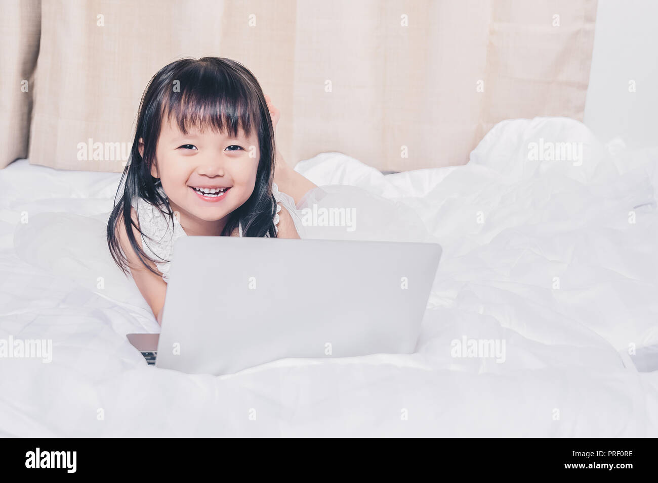 Kleines Mädchen liegt mit Laptop auf dem Bett glücklich lächelnd über das Internet ansehen und Spaß haben, in Beziehung mit Technologie Konzept Stockfoto