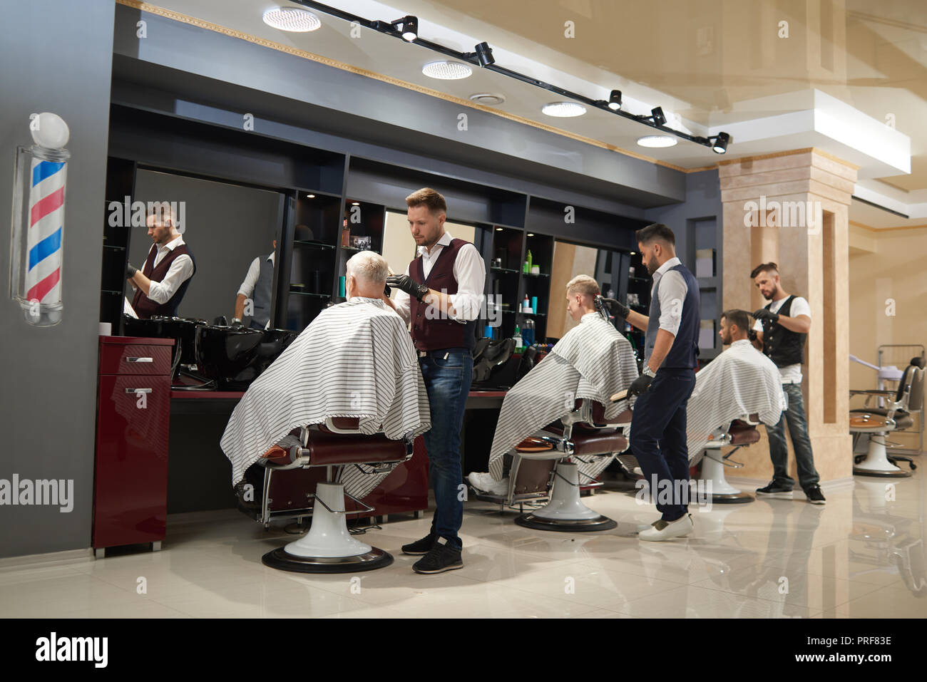 Professionelle Friseure im stilvollen für einheitliches Styling und Pflege Sicherheitsabschläge für Ihre Kunden. Männer sitzen in Stühlen und tragen gestreifte Haarschnitt Kleider. Moderne barbershop Interieur. Stockfoto
