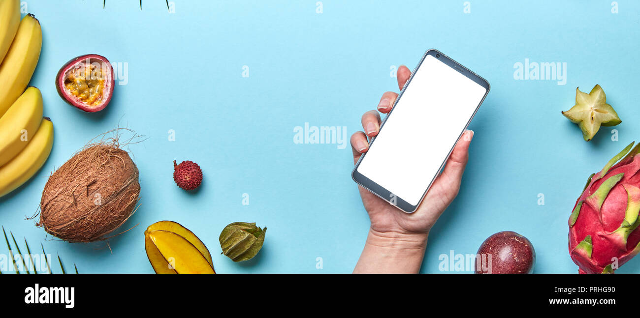 Kokosnuss, Lychee, Ananas Set von tropischen Früchten. Die Hand des Mädchens hält das Telefon auf einem blauen Hintergrund mit einer Kopie des Raumes. Das Konzept der modernen online einkaufen. Flach Stockfoto