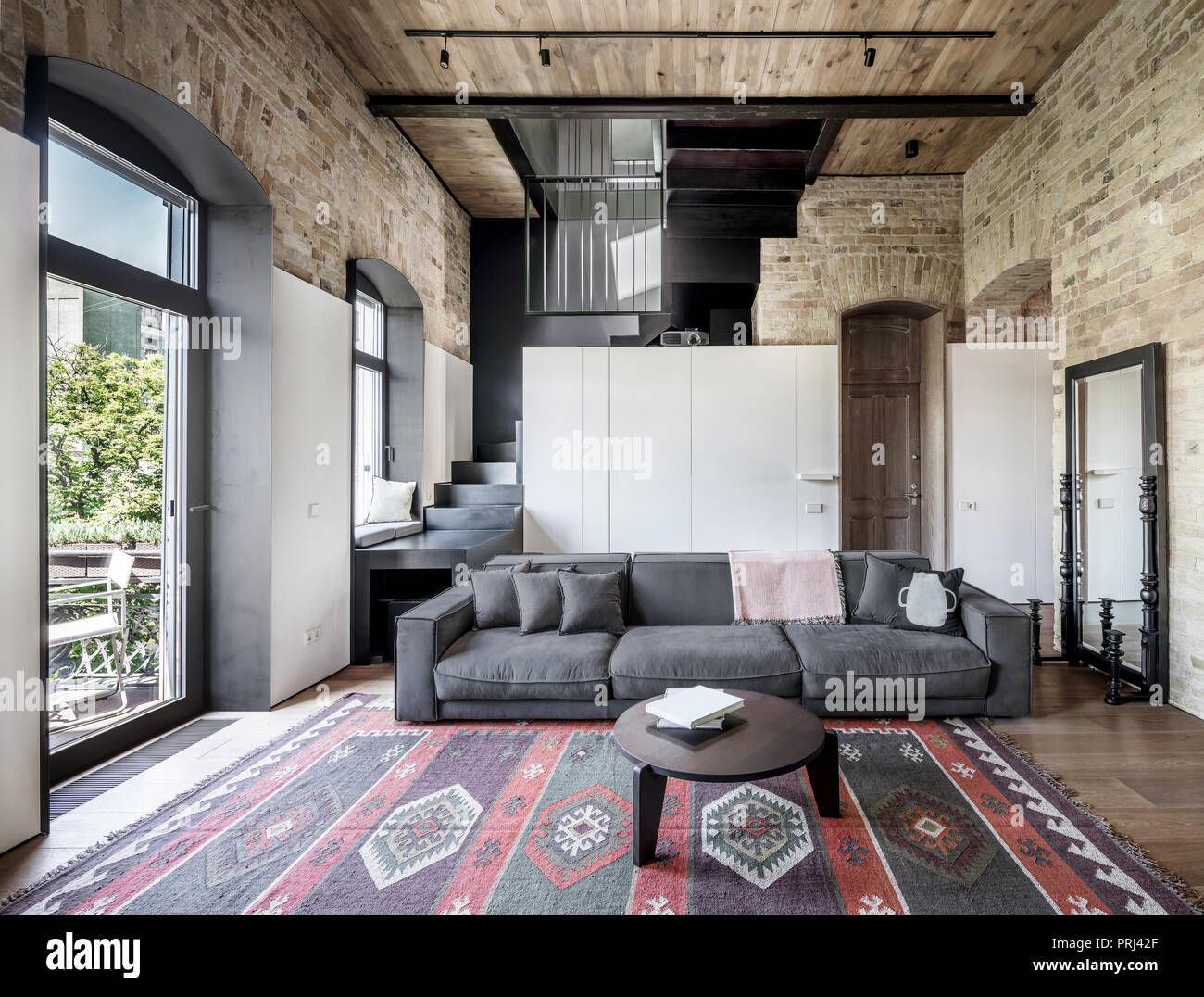 Interieur in einem Loft Stil mit gemauerten Wänden, Holzdecken und ein Parkett mit einem Teppich auf dem Boden. Es ist ein graues Sofa mit Kissen, runder Tisch, st Stockfoto
