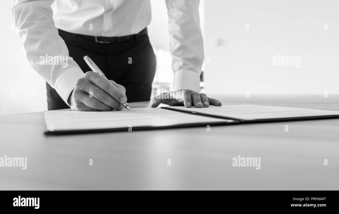 Graustufenbild des Geschäftsmannes, der an seinem Schreibtisch unterzeichnen Vertrag oder juristische Dokument. Stockfoto