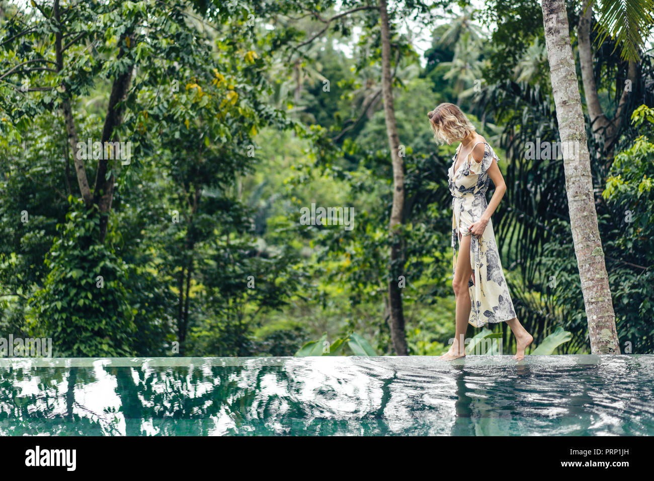 Attraktive blonde Frau im Kleid zu Fuß in der Nähe von Schwimmbad mit grünen Pflanzen auf Hintergrund, Ubud, Bali, Indonesien Stockfoto