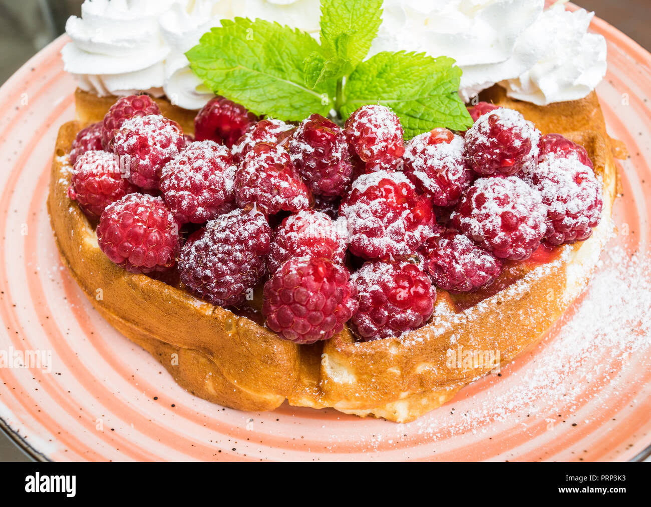 Leckere belgische Waffeln mit frischen Himbeeren und Schlagsahne mit Mandel- Flocken. Mit Minze und Puderzucker eingerichtet, auf orange Platte. Del Stockfoto