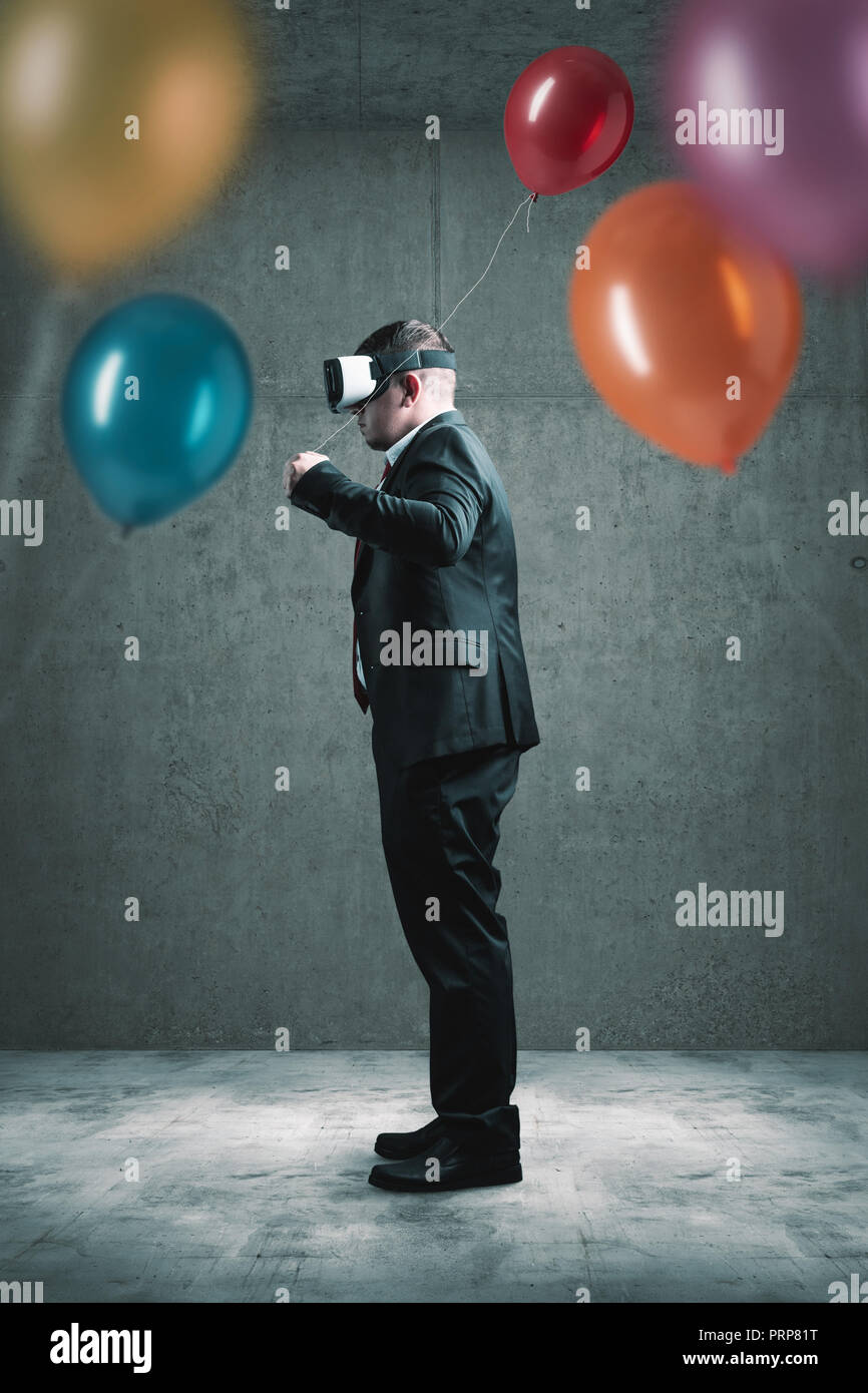 Unternehmer stehen mit VR-Headset auf Betonboden und halten roten Ballon in der Hand vor vielen anderen bunten Ballons Stockfoto